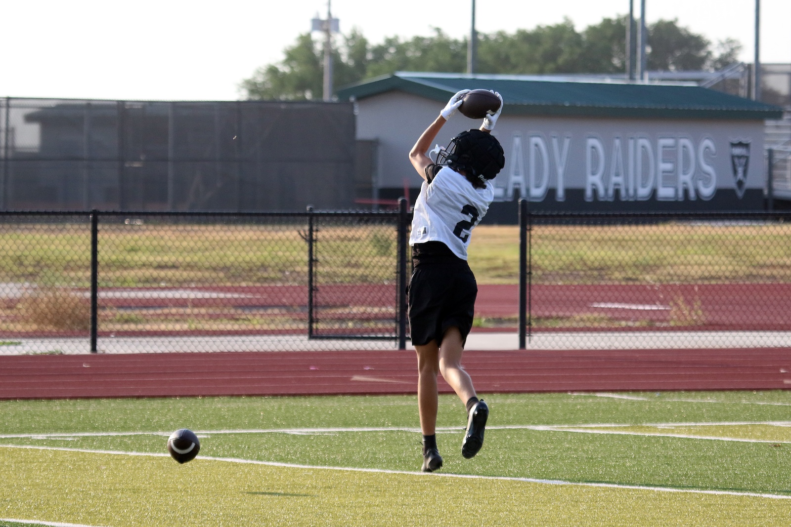 Young football player, white jersey, jumps to catch a ball on a track field with a'Lady Raiders' sign in the background.