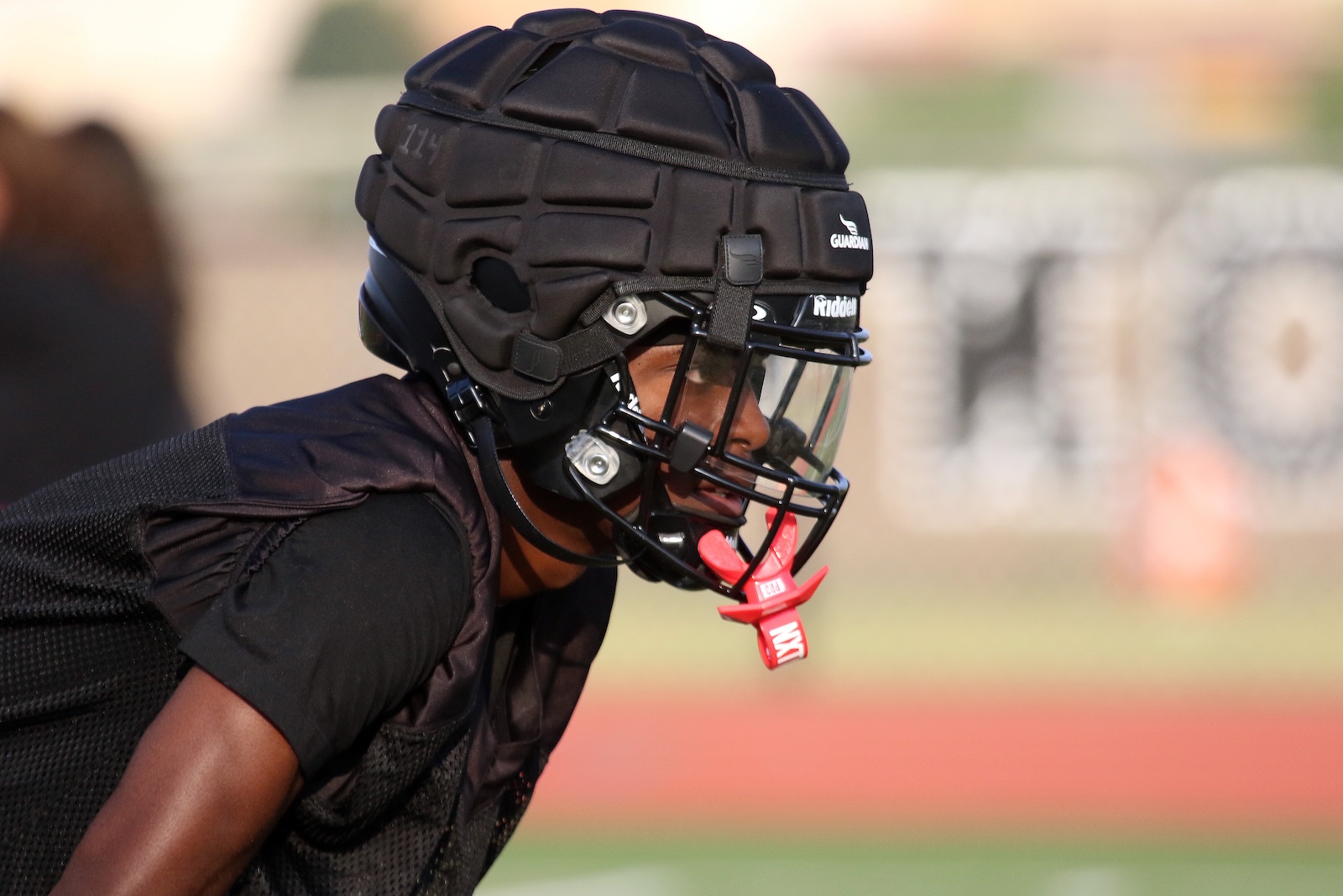 American football player in a black helmet and protective gear crouches on the field, awaiting the snap.