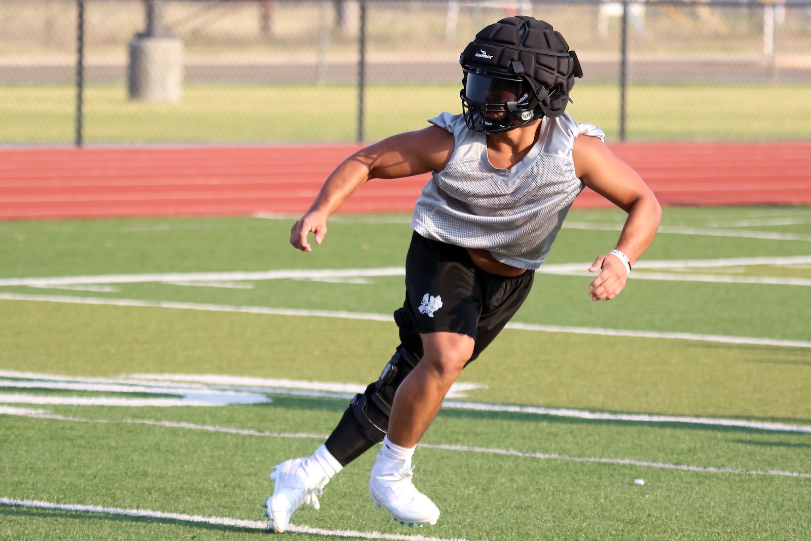 Male football player in a gray sleeveless jersey and black shorts, wearing a protective helmet and knee brace, sprinting on a green turf field.