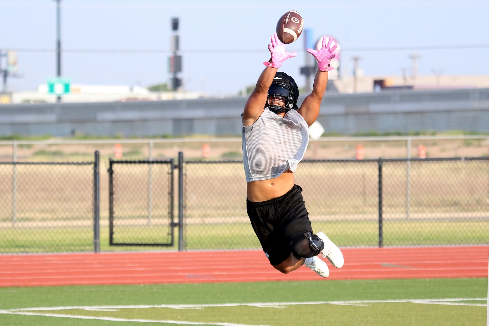 Athlete wearing a helmet leaps to catch a football with pink gloves on a track field fence in the background
