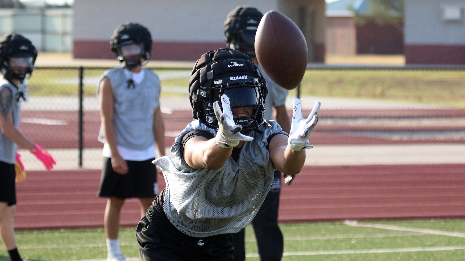 Football player in a helmet and padding reaches for a midair football during practice on a track field.