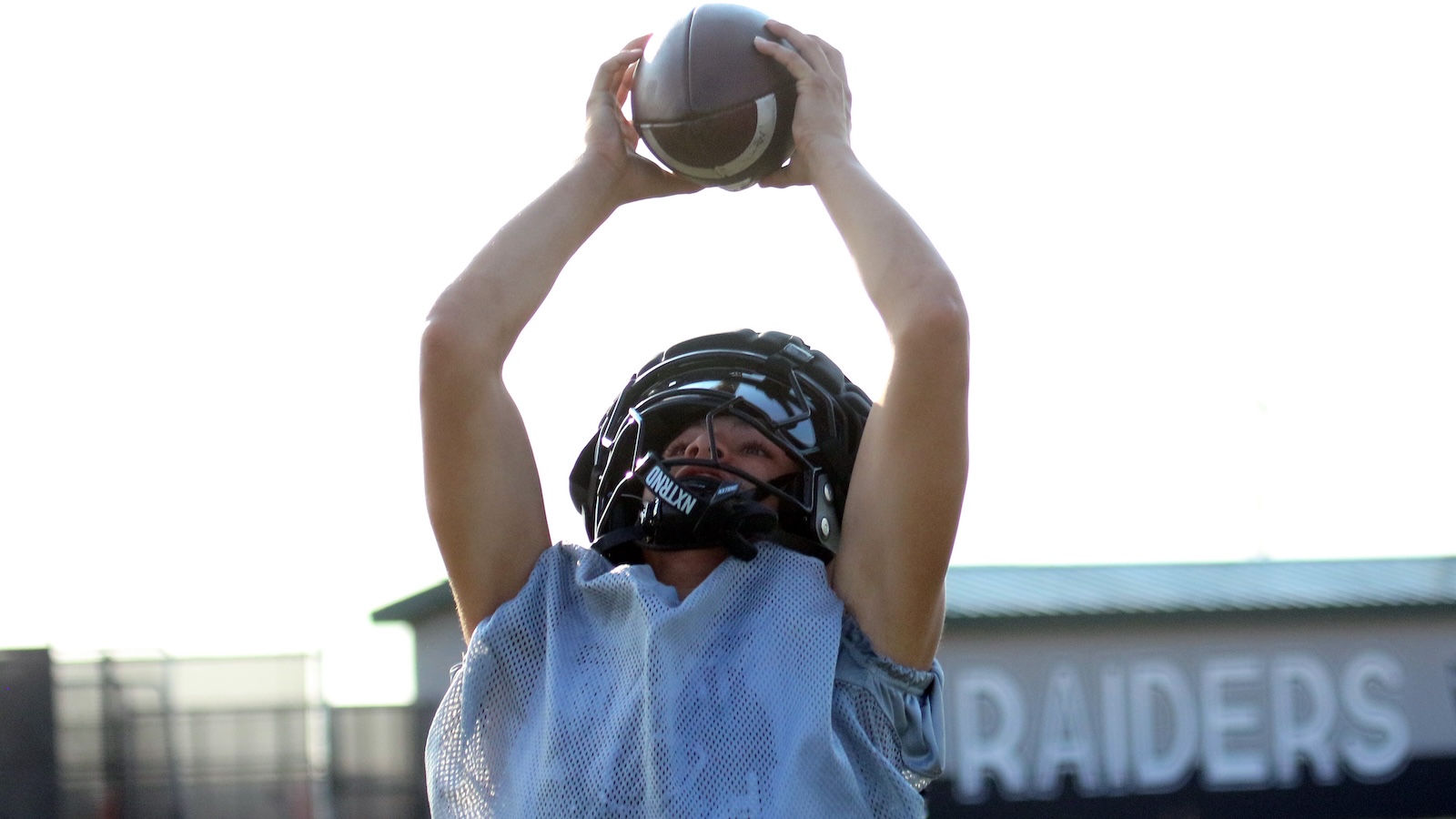 Football player in a helmet and blue jersey reaches upward to catch a ball overhead during drills.