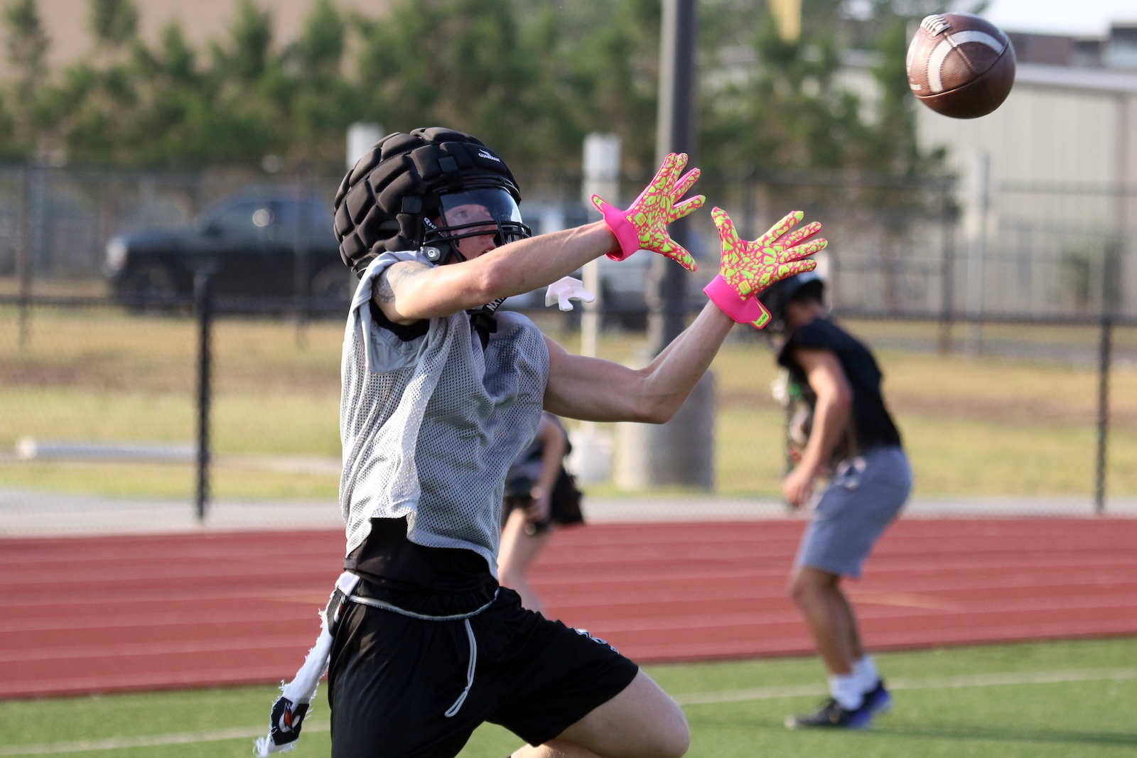 Young football goalie in protective helmet and bright pink gloves stretches to catch a football on a track field outdoor game actio .