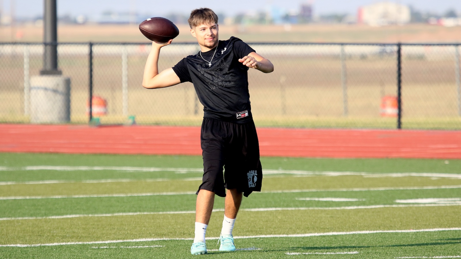 Young man in black athletic wear throwing a football on a turf field with a chain-link fence behind him