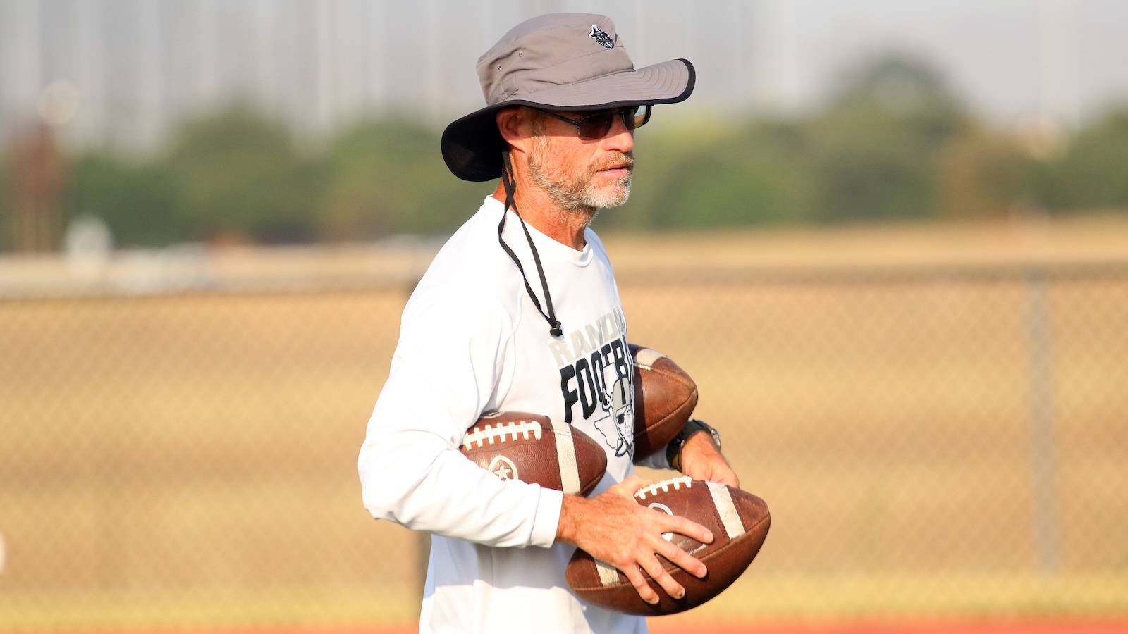 Man with a wide-brim hat and sunglasses holding two footballs on a practice field, ready to throw or coach.