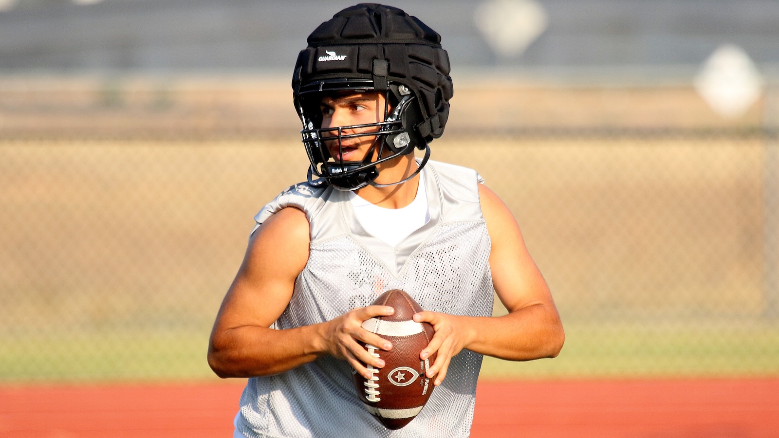 Young football quarterback in a black helmet and gray practice jersey grips a brown football on a field.
