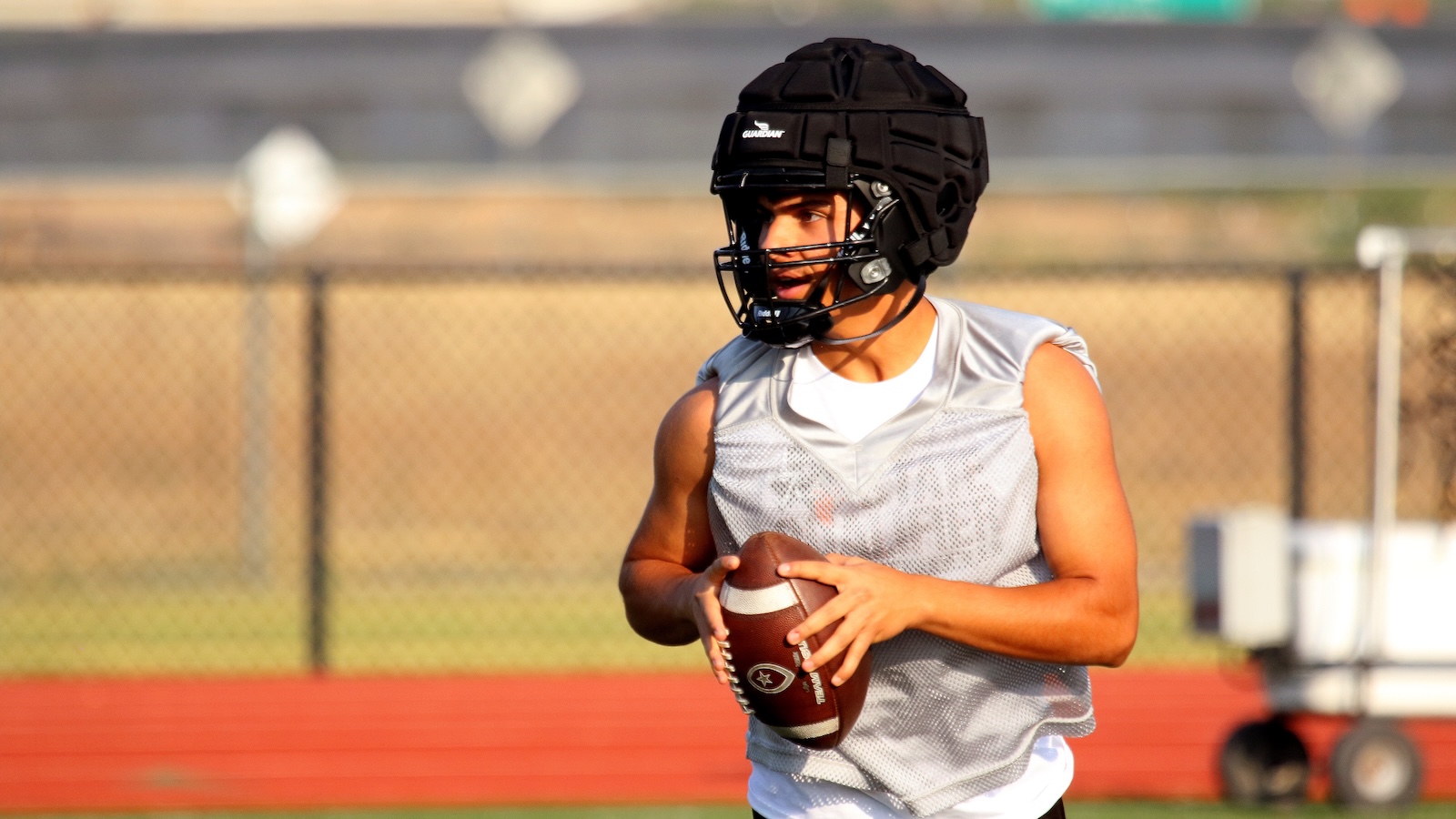Football player wearing a protective helmet and mesh practice jersey holds a ball, ready to throw on a sunny field near a fence.