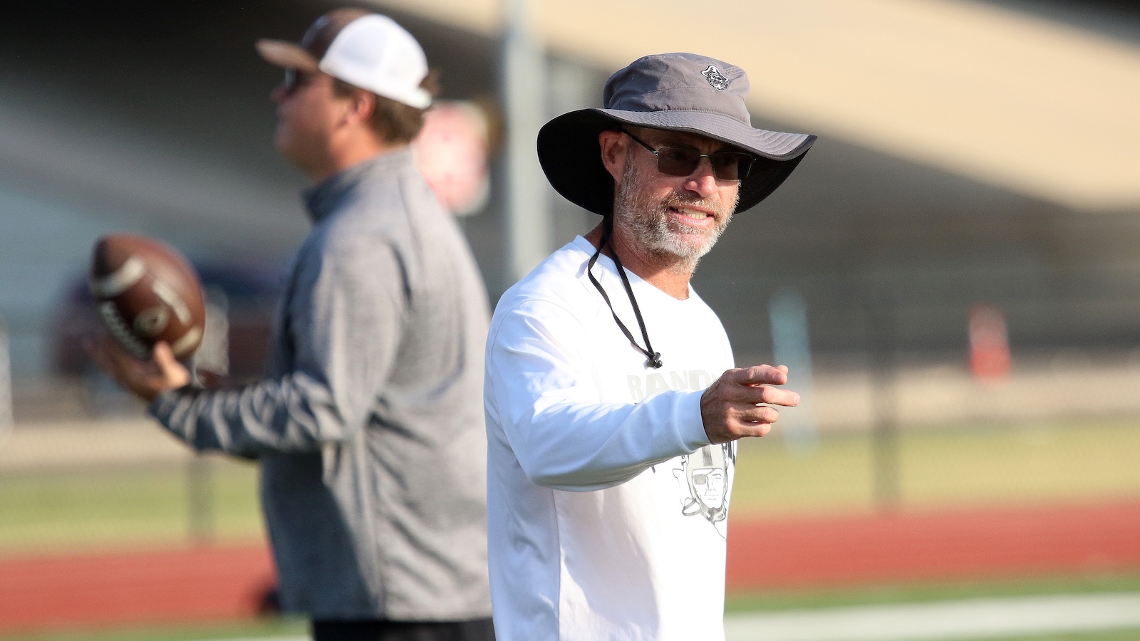 Coach in a white shirt and wide-brim hat points while talking on a football field; another person with a football stands in the background.