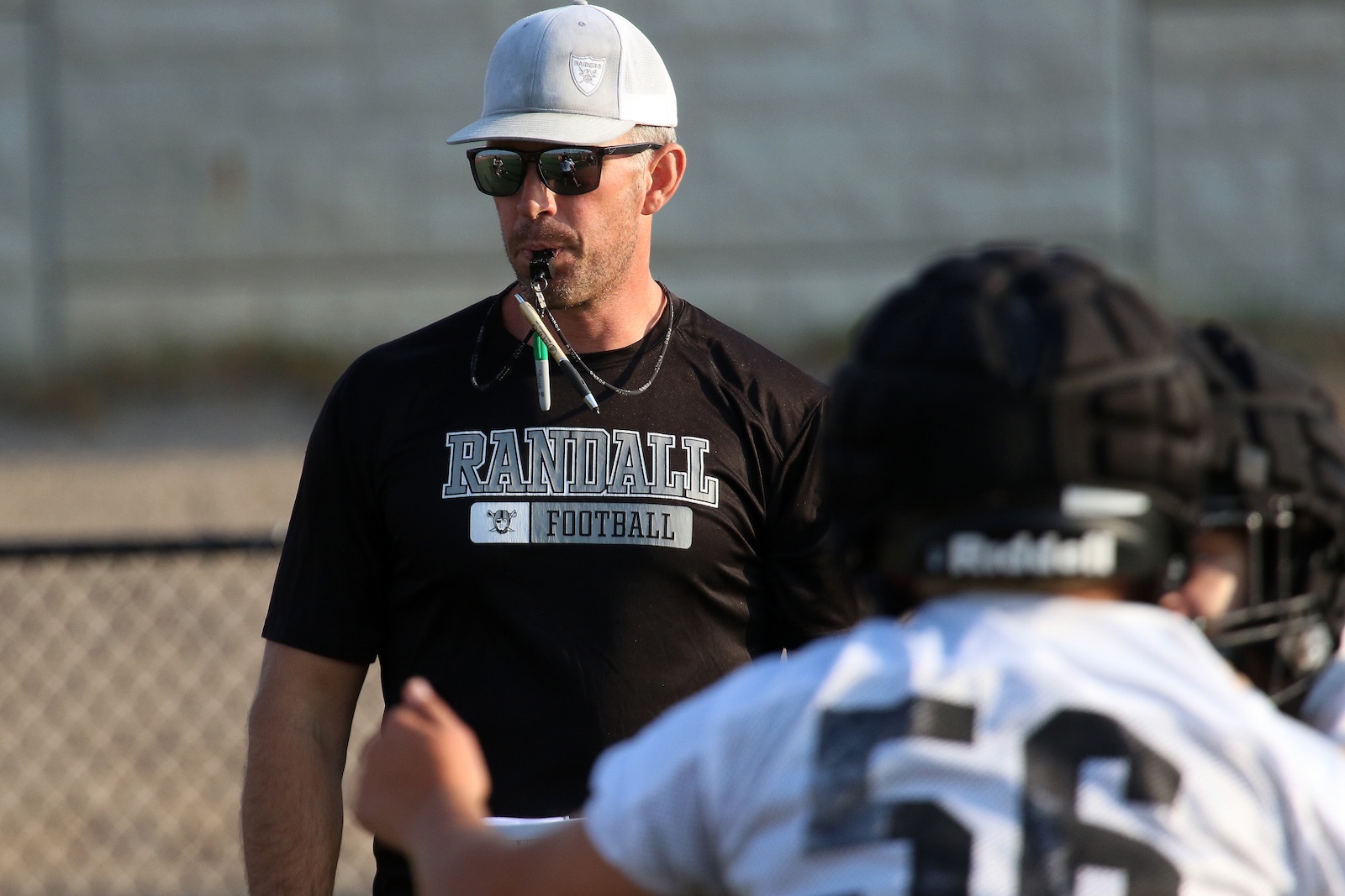 Coach in a white cap and sunglasses watches players on a football field; ‘Randall Football’ shirt is visible.