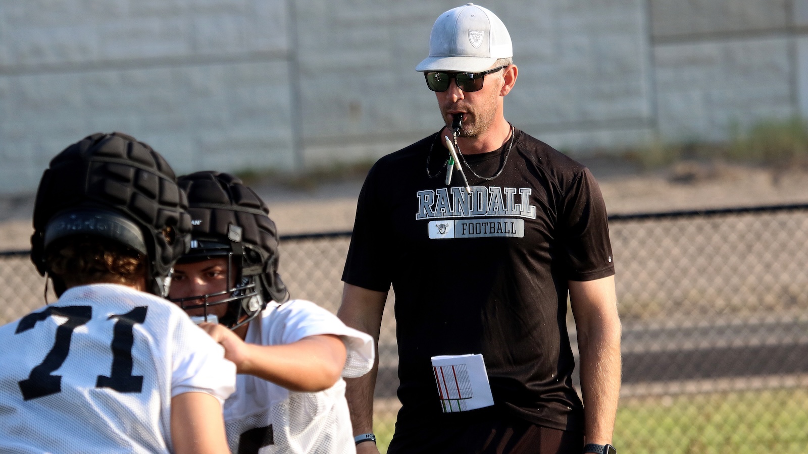 Football coach in a white cap and sunglasses supervising two players during practice on a football field.