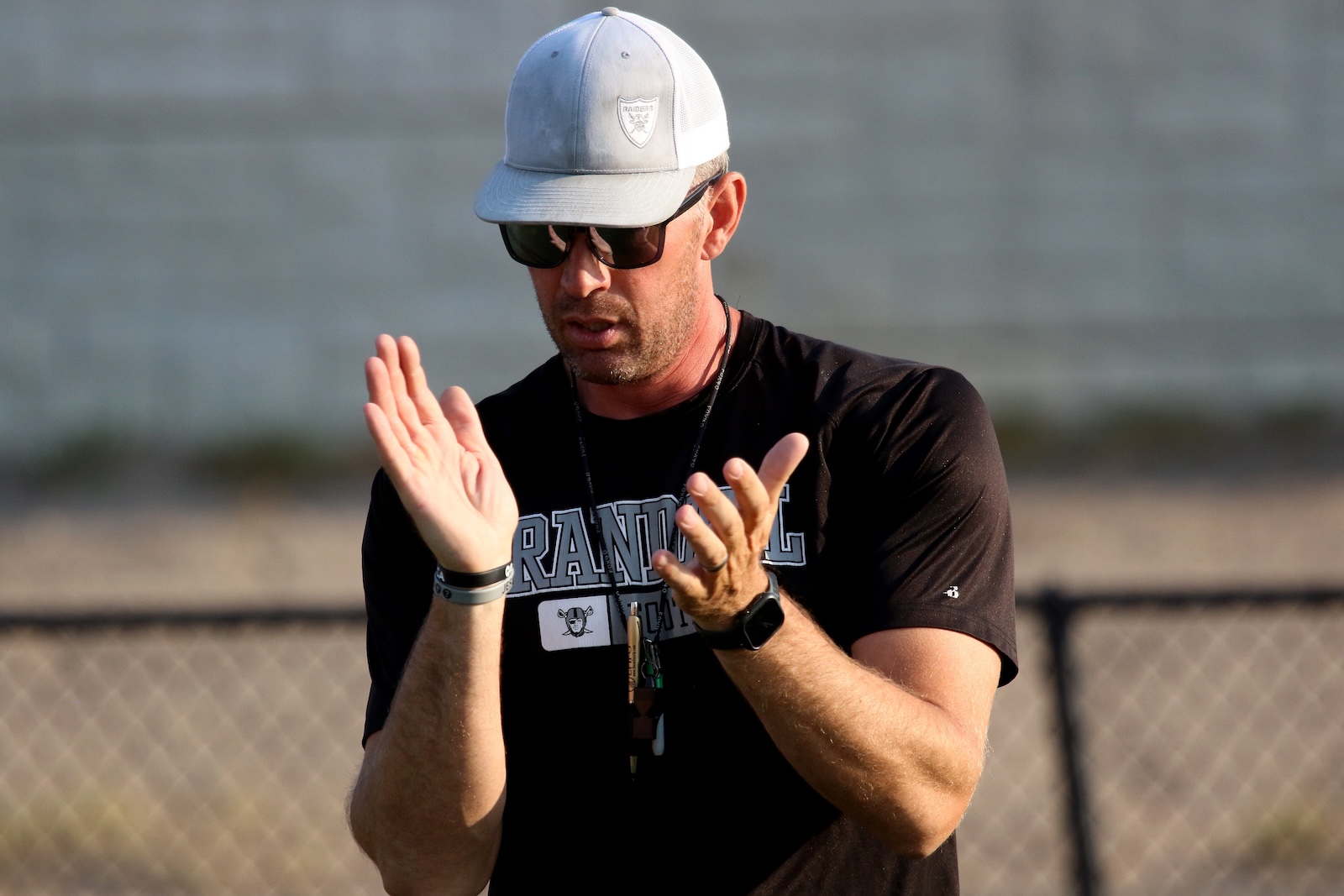 Man wearing a gray cap and sunglasses claps with raised hands outdoors near a chain-link fence.