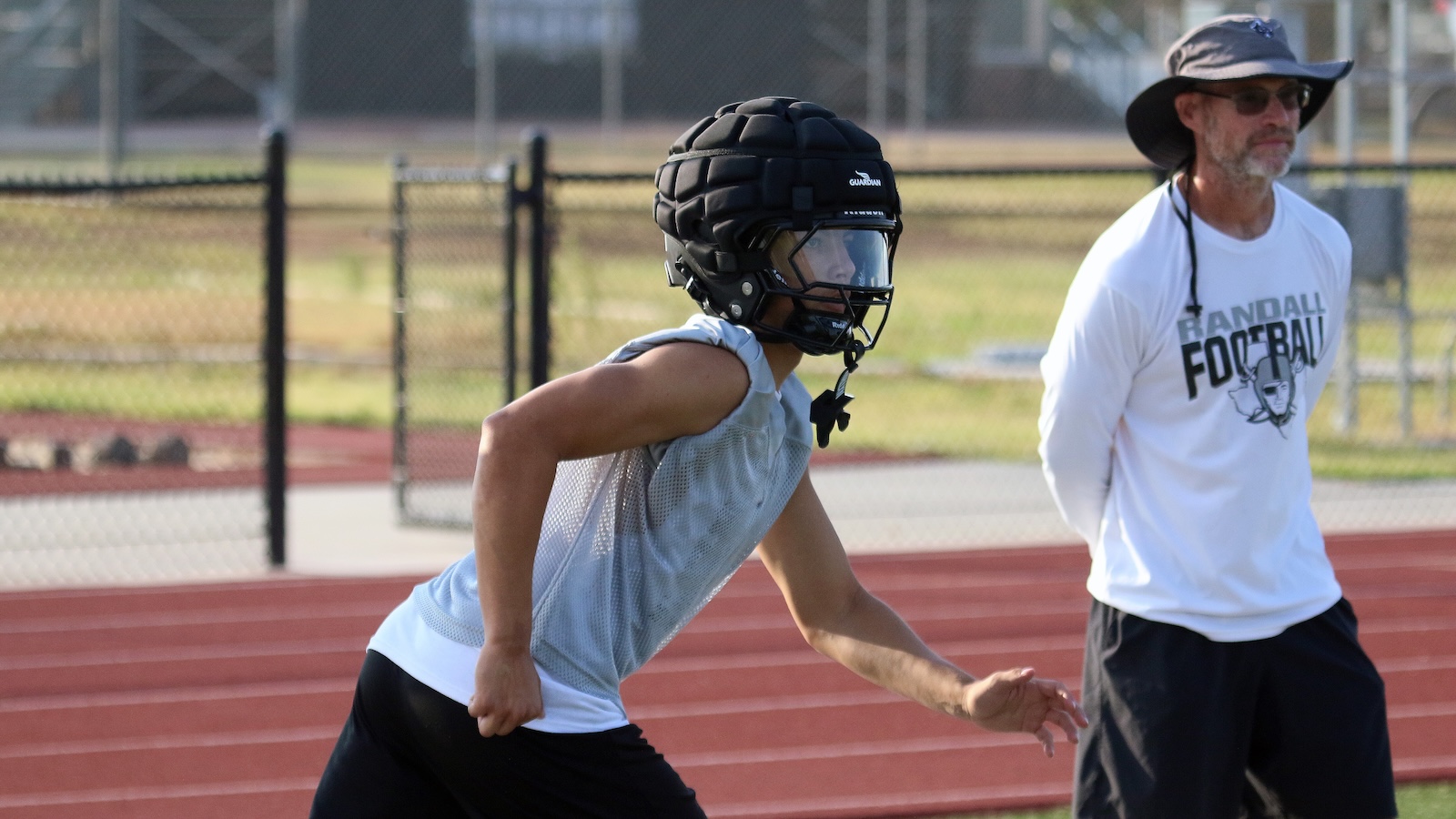 Young football player wearing a black protective helmet and gray sleeveless shirt crouches on a track while a coach in a white Randell Football shirt watches nearby.