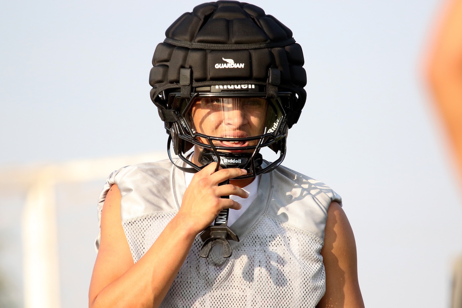 Person wearing a black padded football helmet with a face guard, holding a mouthguard against the chin, outdoors in a gray mesh jersey.