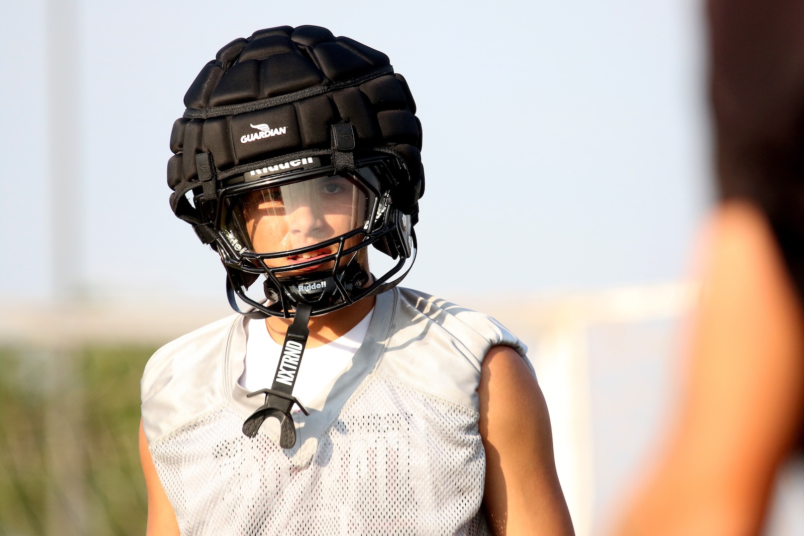 Athlete wearing a black protective helmet with faceguard and a gray mesh jersey, looking toward the camera outdoors.
