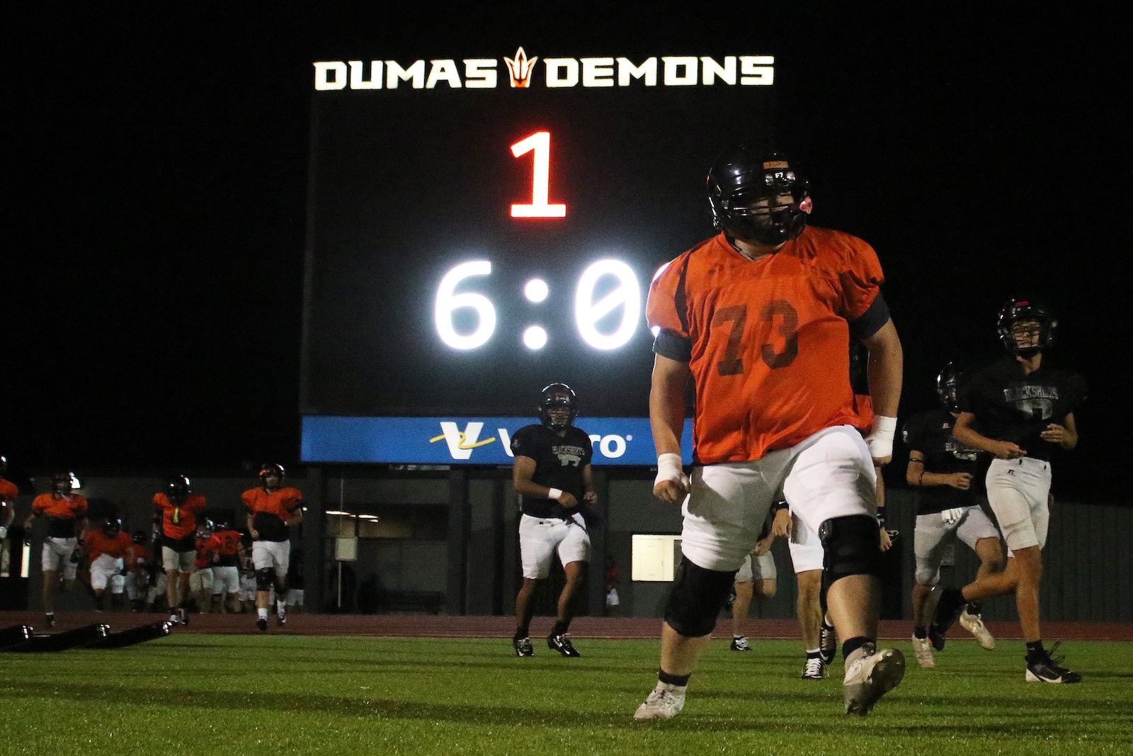 Nighttime football scene: players in orange jerseys walk onto the field as a lit scoreboard reads DUMAS DEMONS with a 6:0 score.