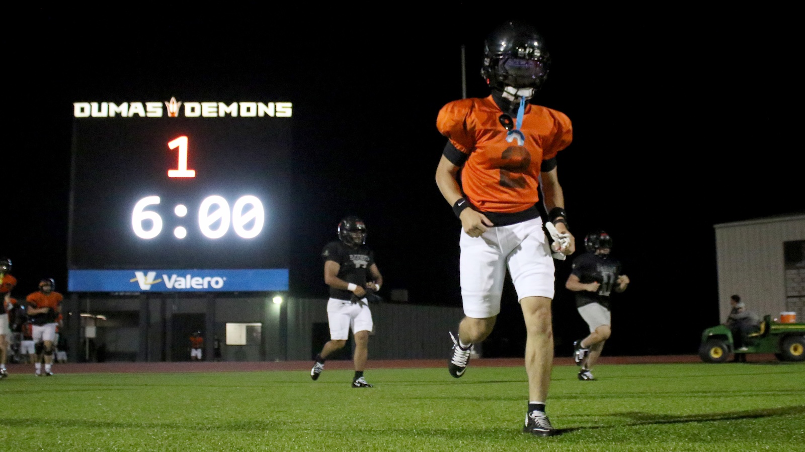 Nighttime football field with players in orange and black; scoreboard reads'DUMAS DEMONS' and 6:00 remaining.