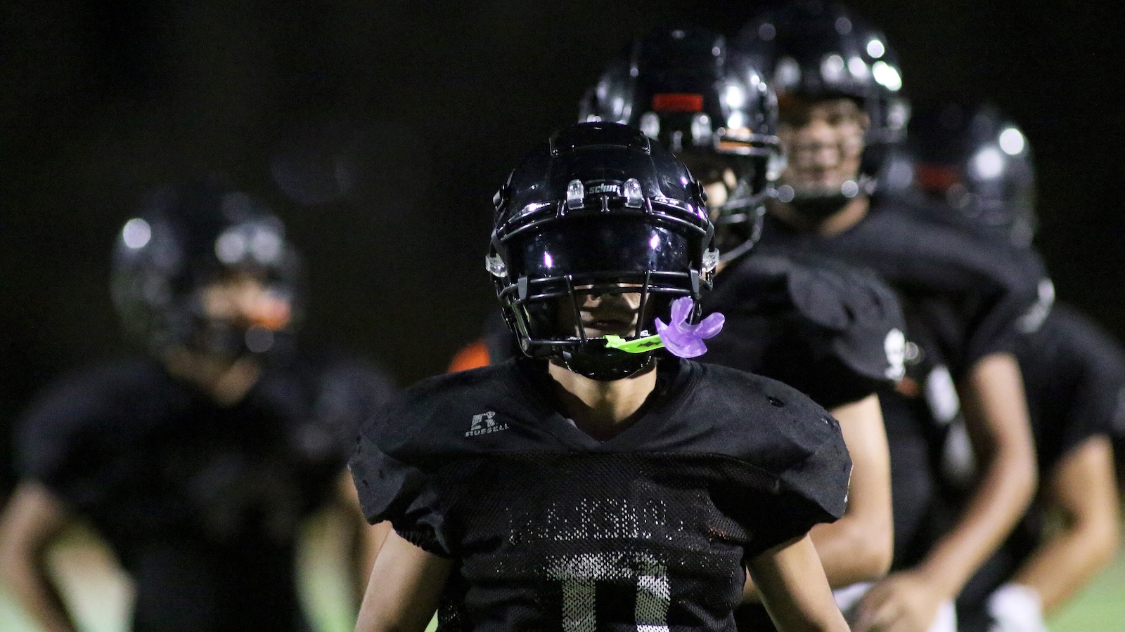 Nighttime football practice with players in black uniforms; front player wears a helmet and a purple flower on his mouthguard.