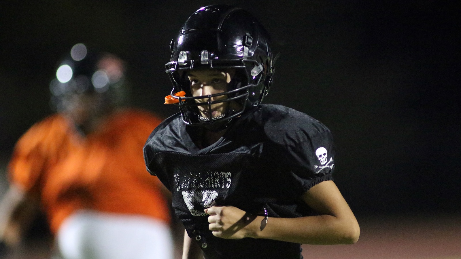 Young football player in a black helmet and jersey running on a dimly lit field at night, wearing a skull emblem on the sleeve UIO helmet chin strap visible.
