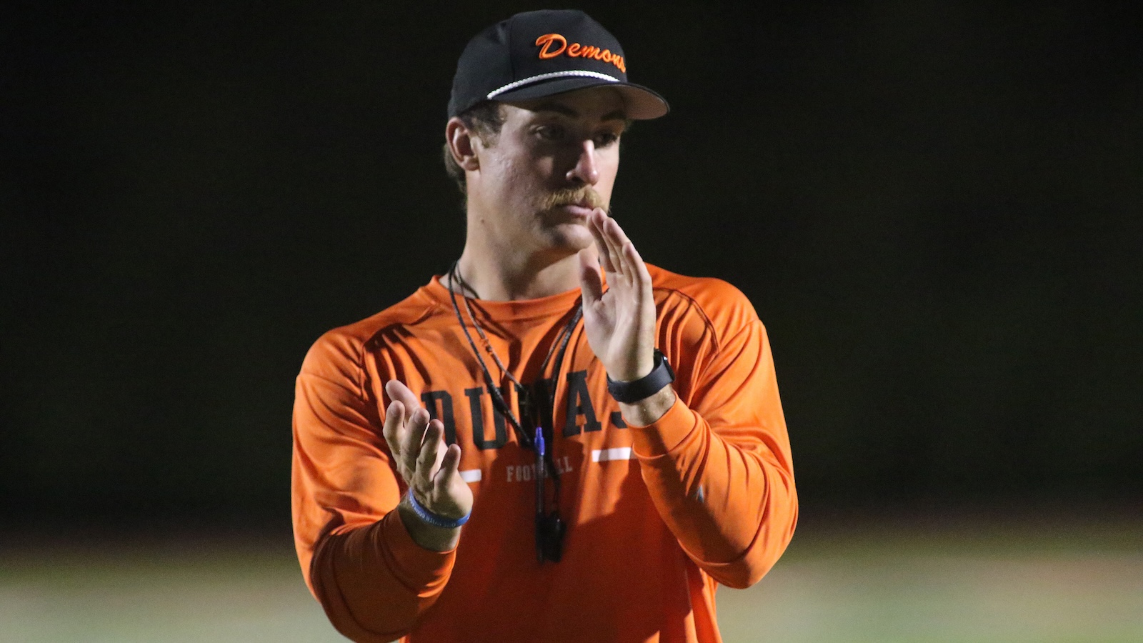 Male coach in an orange long-sleeve shirt claps on a football field at night, wearing a cap and whistle lanyard nearby.