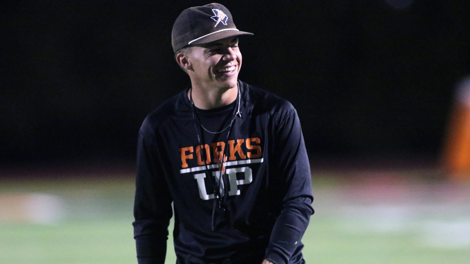 Smiling man in a dark long-sleeve shirt with'FORKS UP' on the chest, wearing a cap, on a football field at night (coaching/athlete vibe).