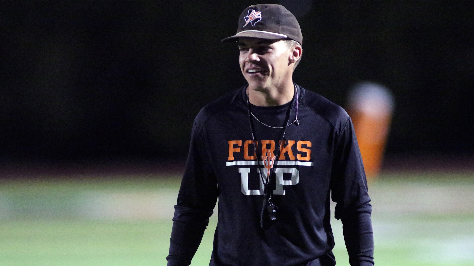 Young man in a black'FORKS UP' shirt and cap on a nighttime practice field, smiling with a whistle around his neck.”,