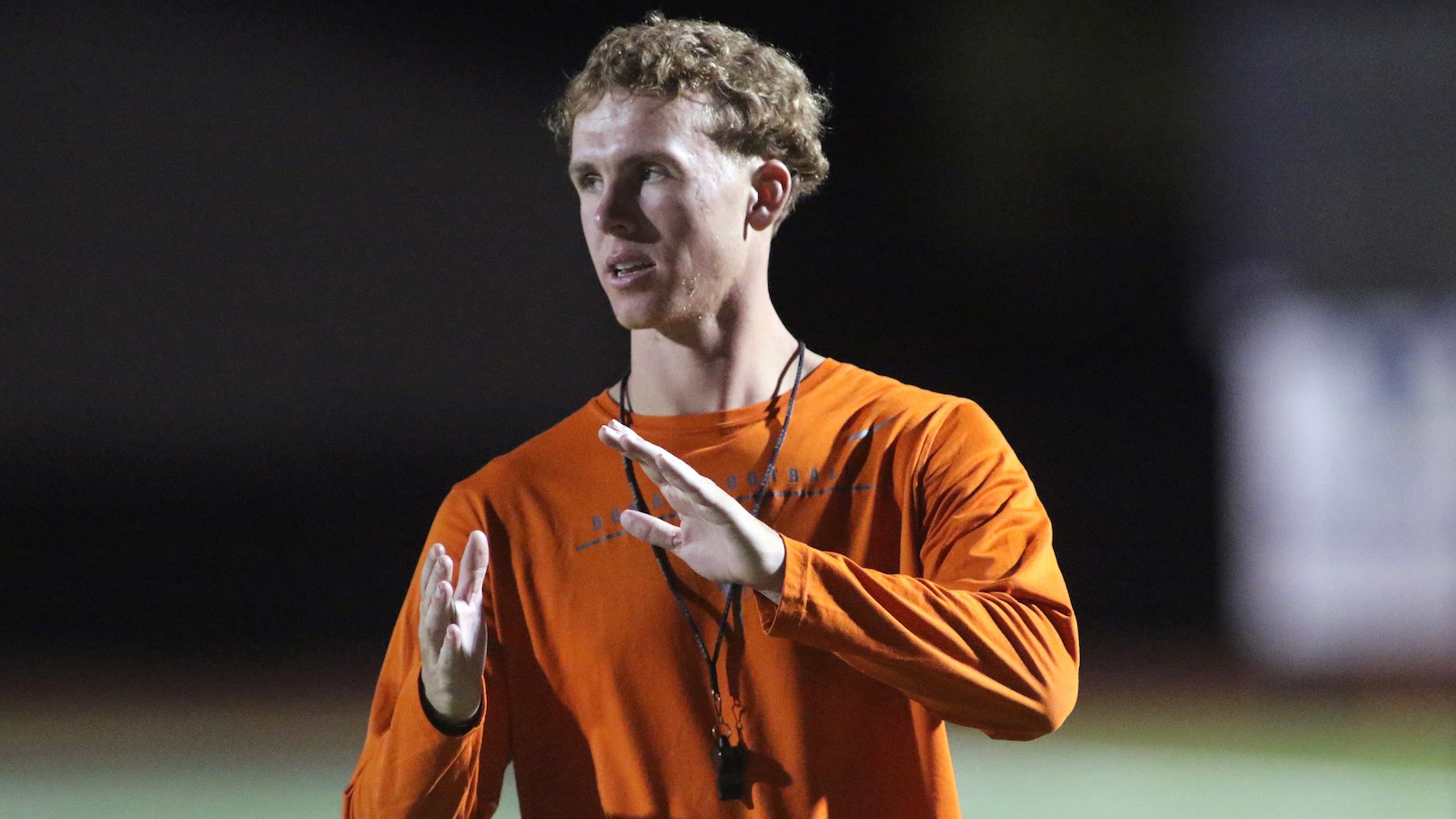 Coach wearing an orange shirt gesturing with his hands while speaking on a sports field at night.