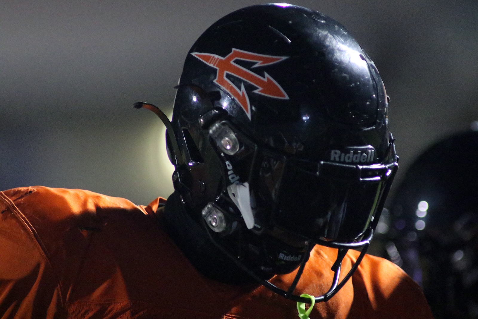 Close-up of a football player wearing a black helmet with an orange stylized'R' logo and an orange jersey at night.