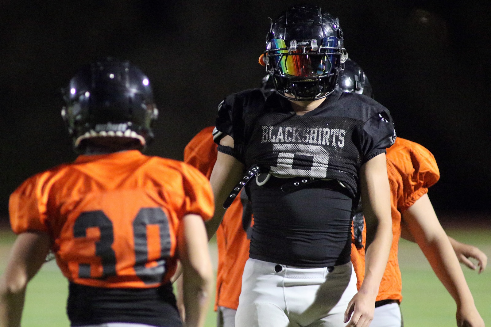 Nighttime football scene with a player in a black'BLACKSHIRTS' jersey facing orange-jersey teammates, wearing a dark helmet with a reflective visor.