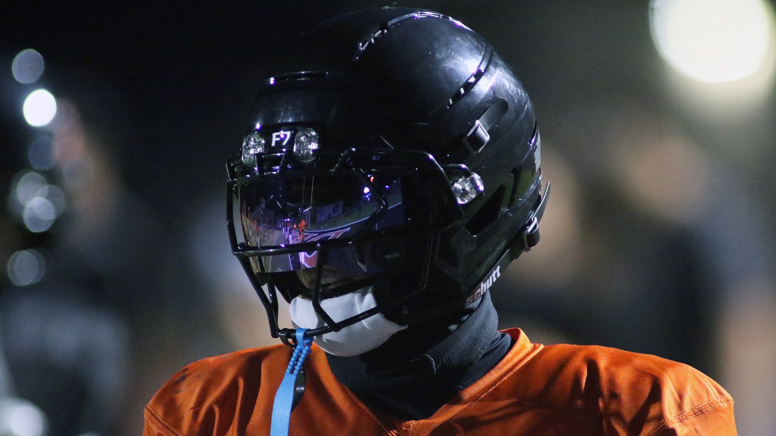 Football player in a black helmet with a tinted visor and orange jersey, facing left in a night game.