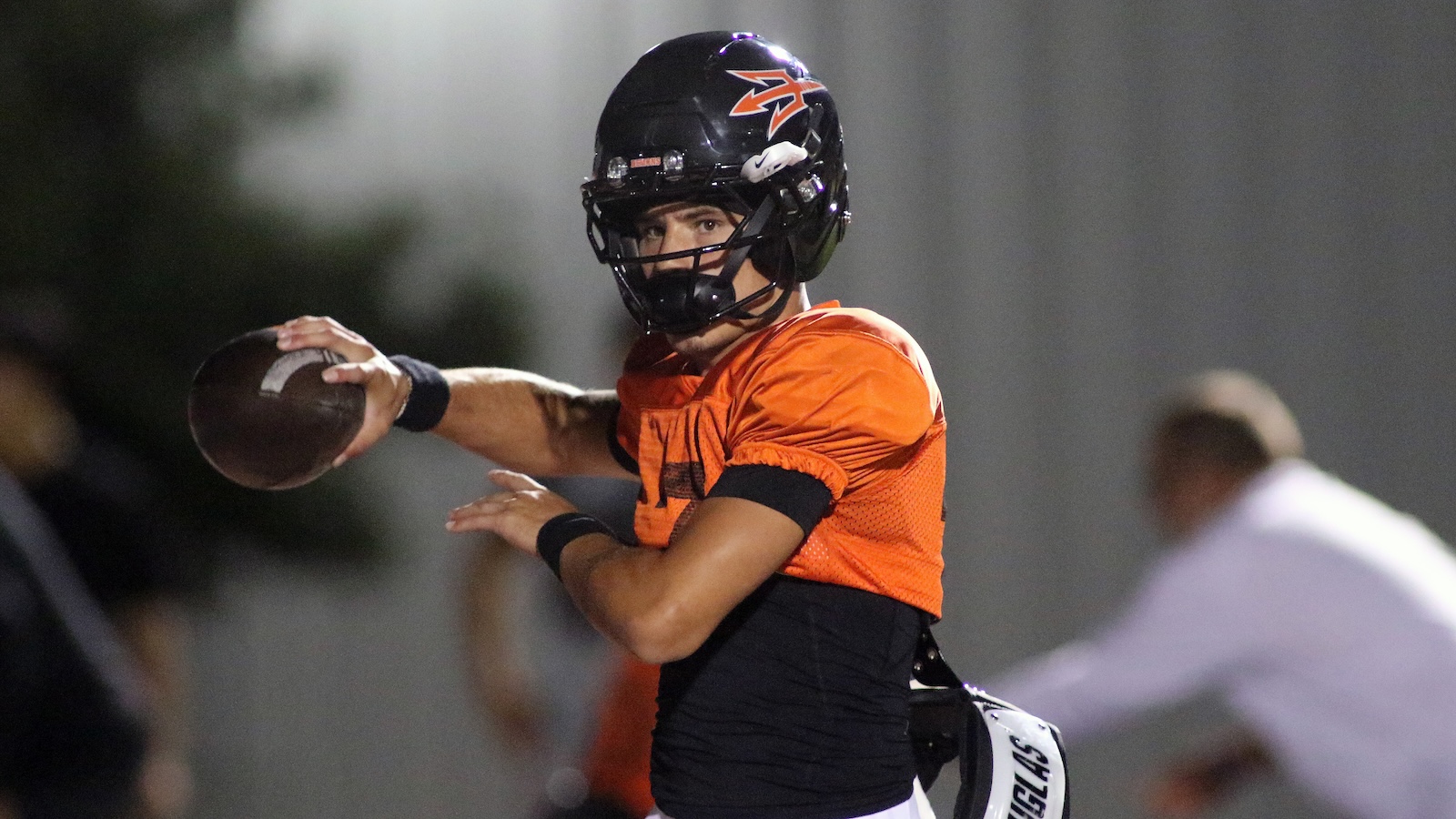 Quarterback in an orange practice jersey winding up to throw a forward pass, wearing a black helmet with a logo.