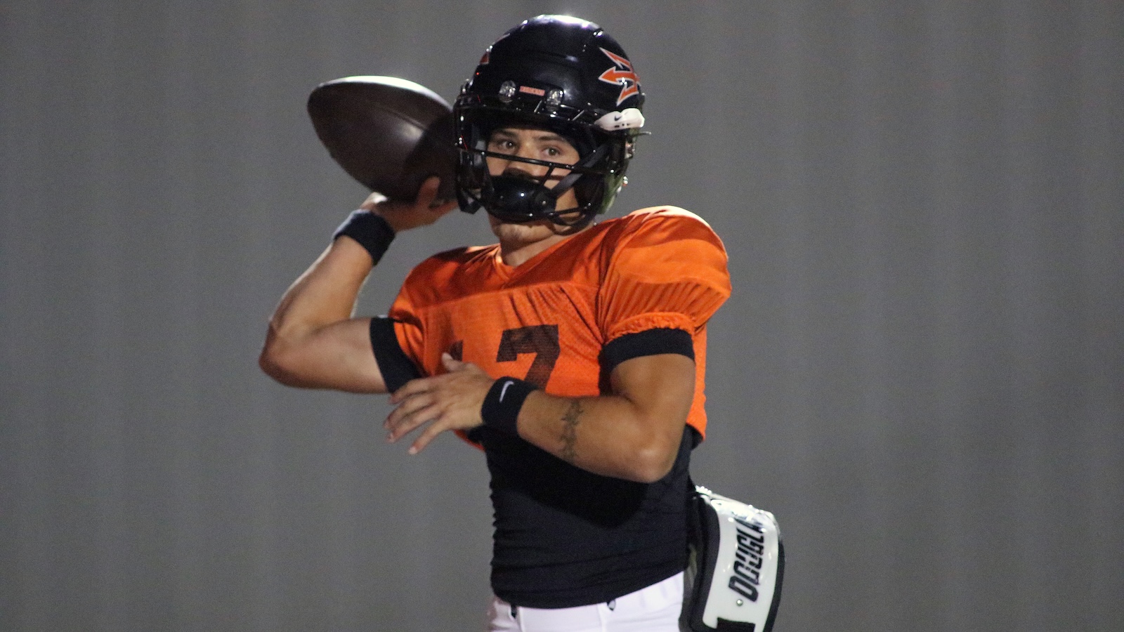 Quarterback in an orange jersey throwing a football mid‑throw, wearing a black helmet and wristbands on a gray background.