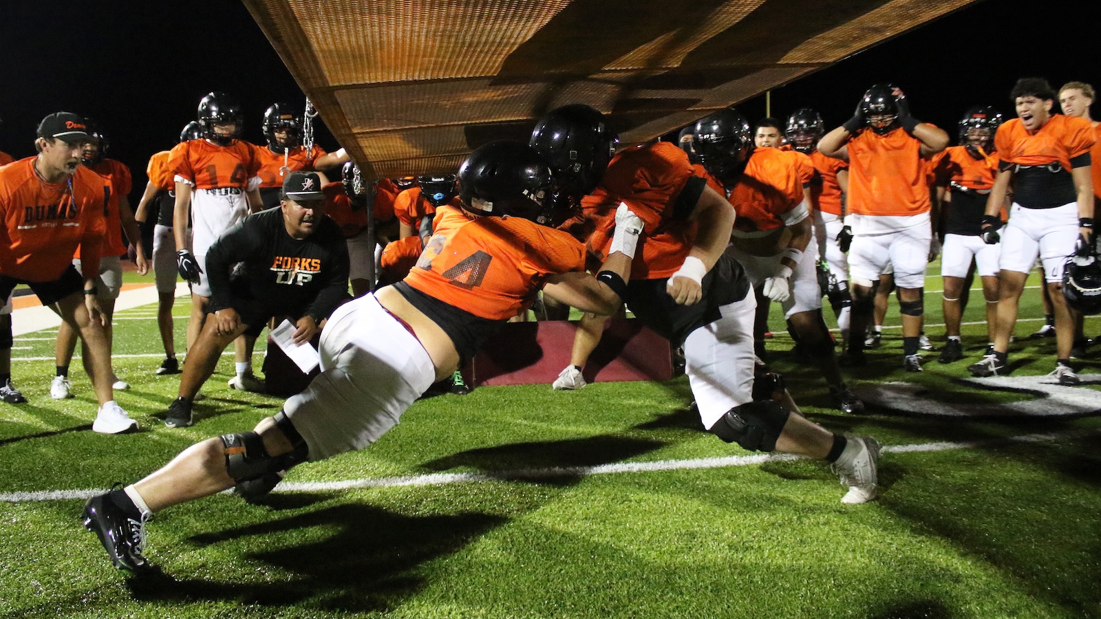 Two football players in orange jerseys dive through a barrier while teammates watch during a night practice on a turf field.