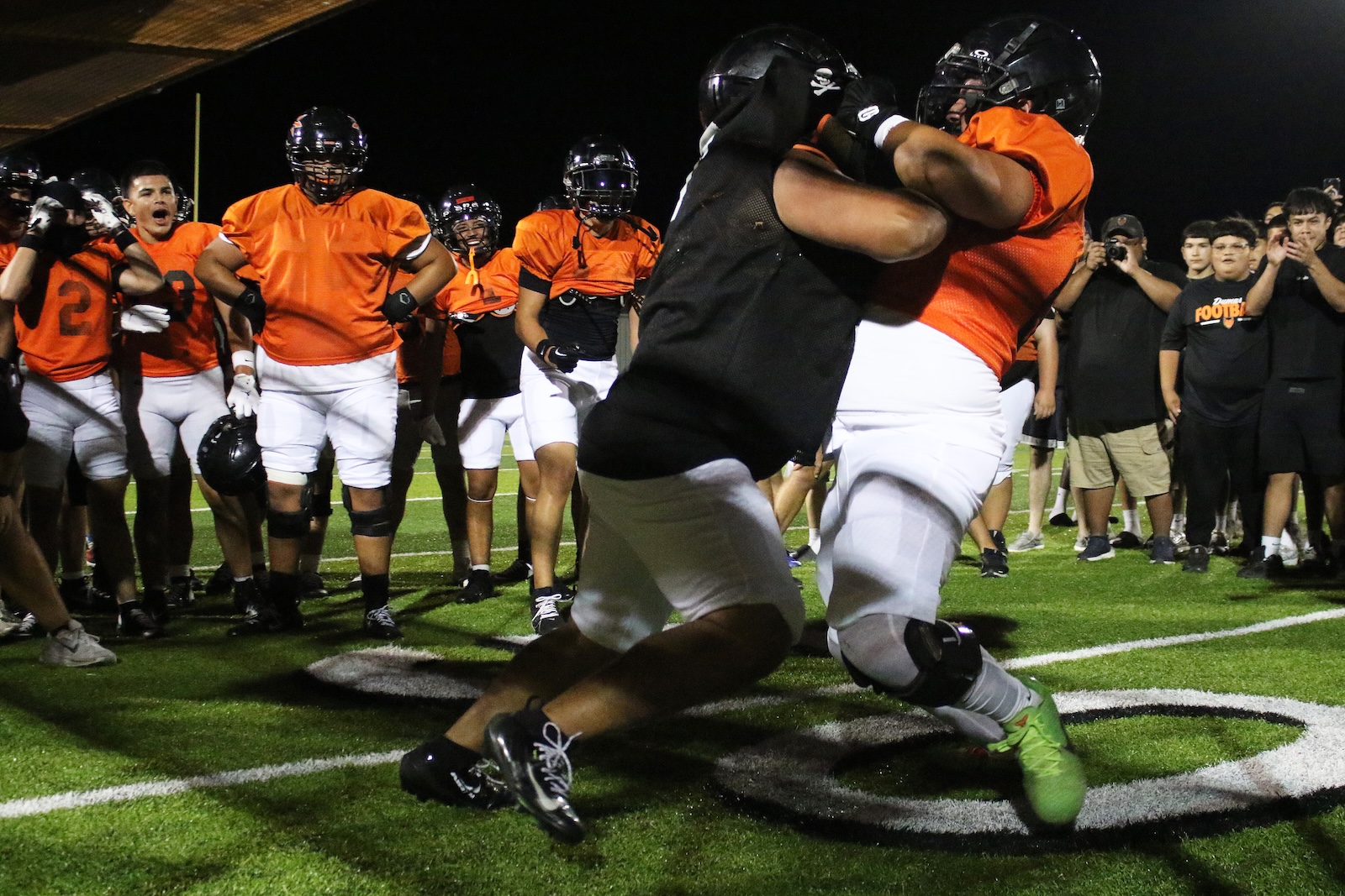 Two football players in orange jerseys grapple in a nighttime drill on a lit turf field, with teammates watching in the background.
