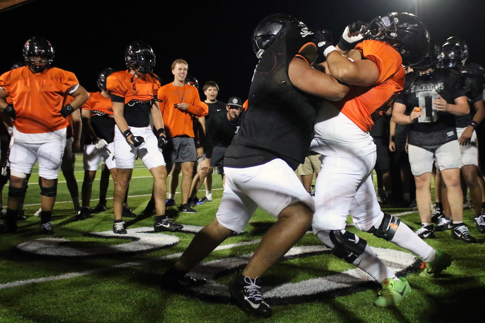 Two football players clash in a night practice, one in black and the other in orange, while teammates watch nearby on the field.