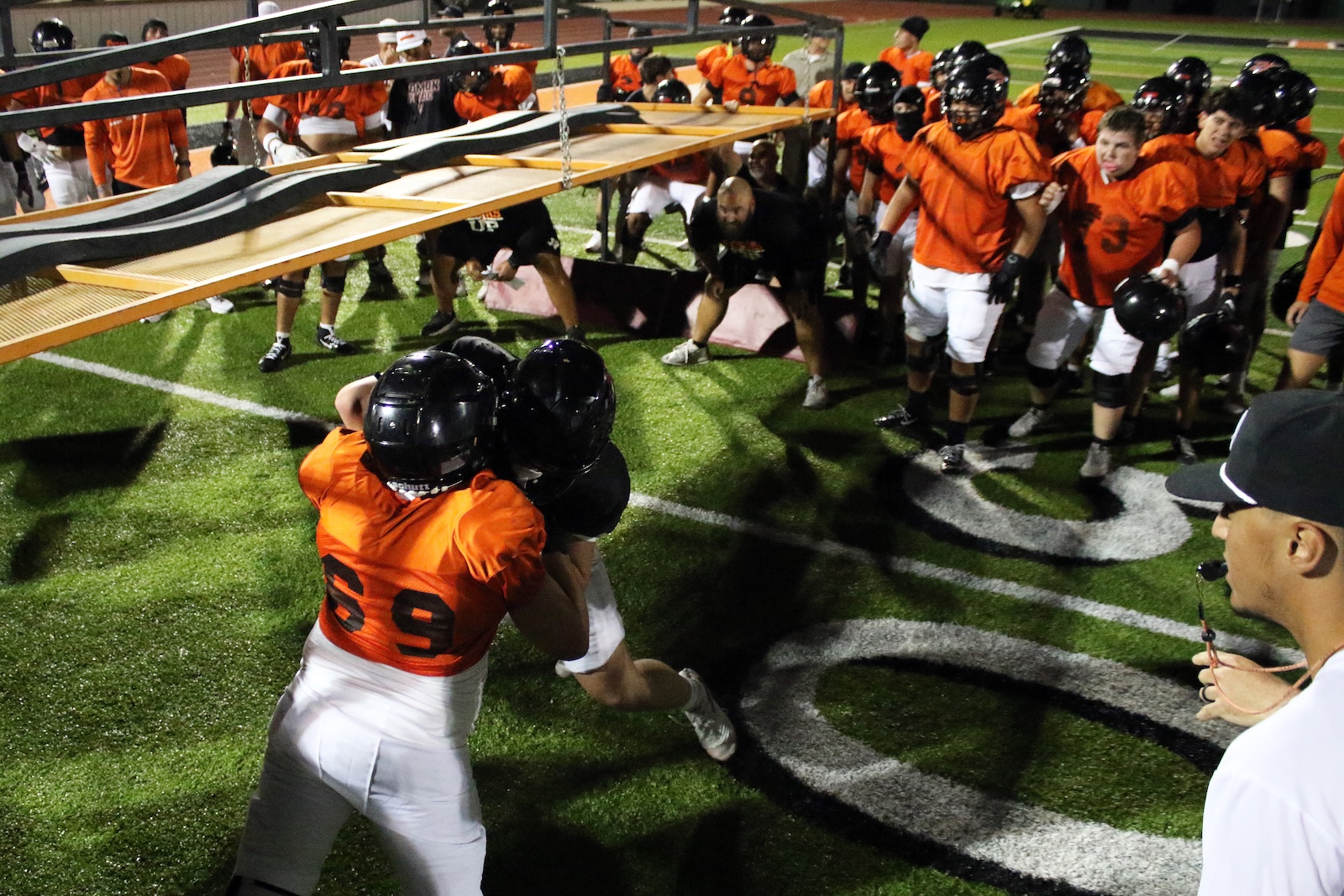 Youth football team in orange jerseys and black helmets hoists a long banner frame over a green turf field at night, surrounded by teammates.