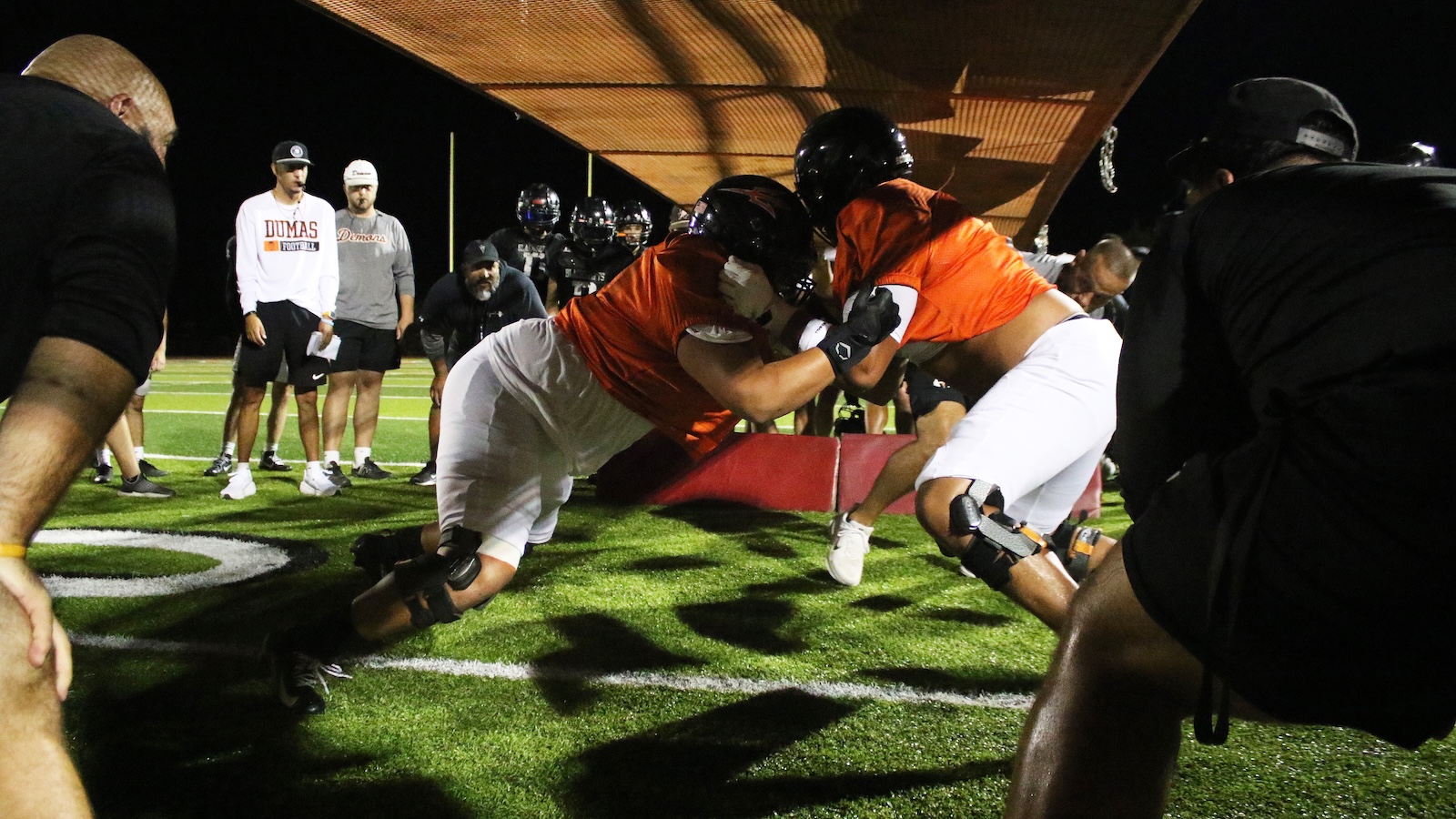 Two football players in orange jerseys collide while pushing against a red blocking pad on a lit night practice field, with teammates watching in the background.