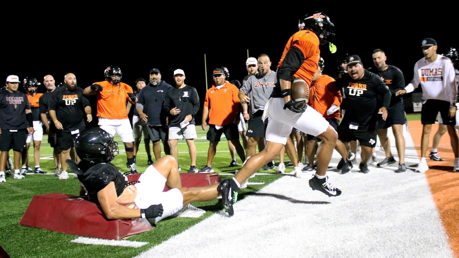 Athletes watch a football drill at night as a player in orange jumps over a tackling pad while another player lies on the pad on the field edge, surrounded by spectators.