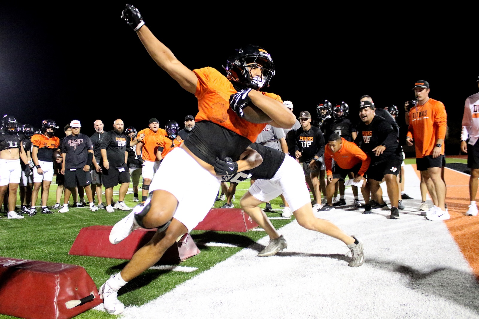 Two football players in orange jerseys collide during a night drill on a turf field, with a crowd of teammates and coaches watching.