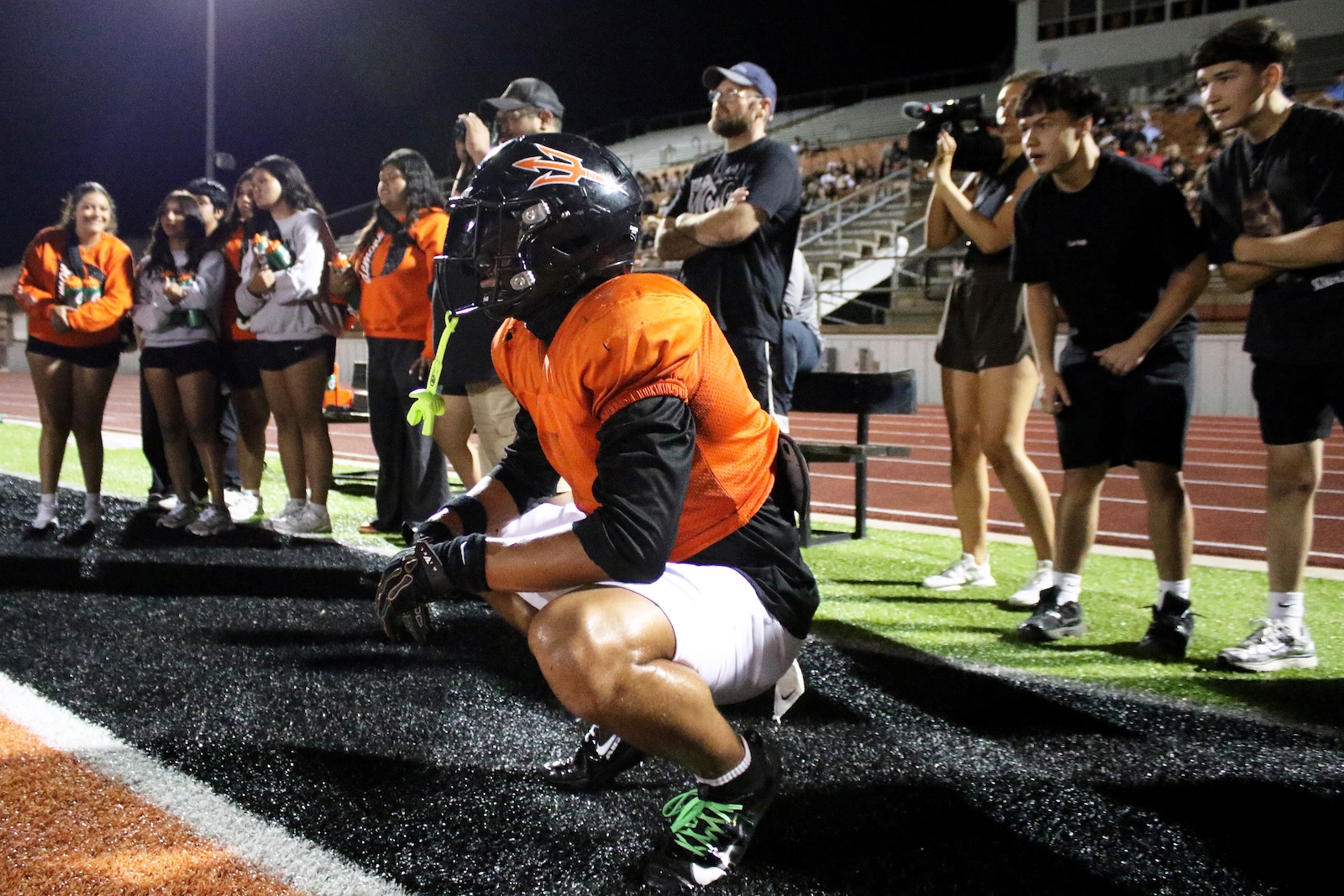 Football player in an orange jersey kneels on turf near the sidelines, crowd watching in the background at night stadium show of support.