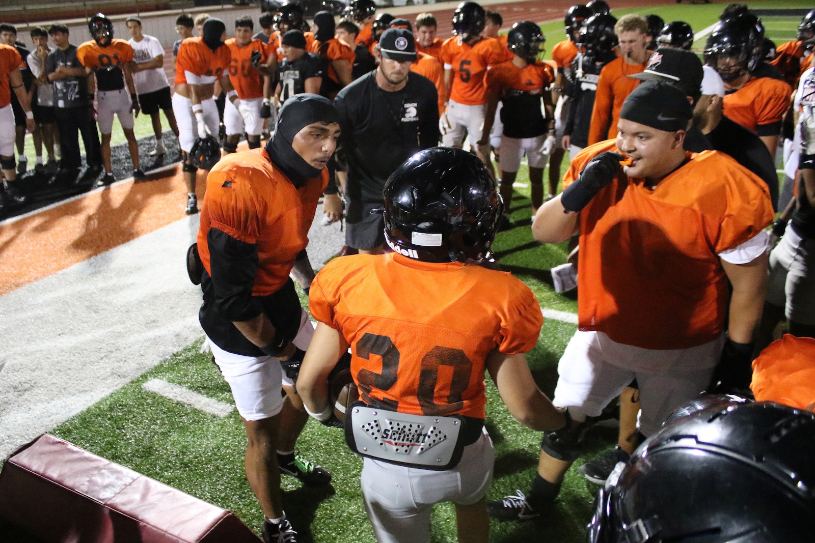 Football players in orange jerseys form a huddle on a synthetic turf field at night, listening to a coach amid a team gathering.