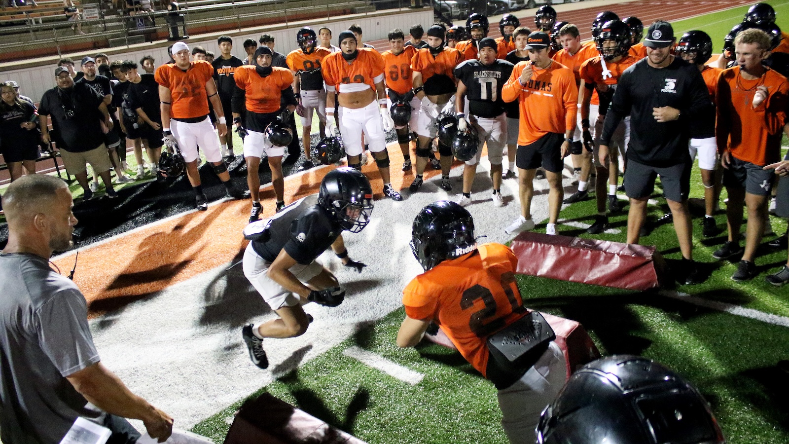 Football practice scene: players in orange and black surround two players in a drill on artificial turf near mats and a coach observing.