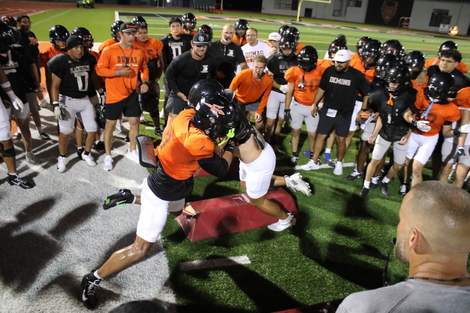 Two football players collide during a drill on the turf while teammates form a circle around them, watching intently.