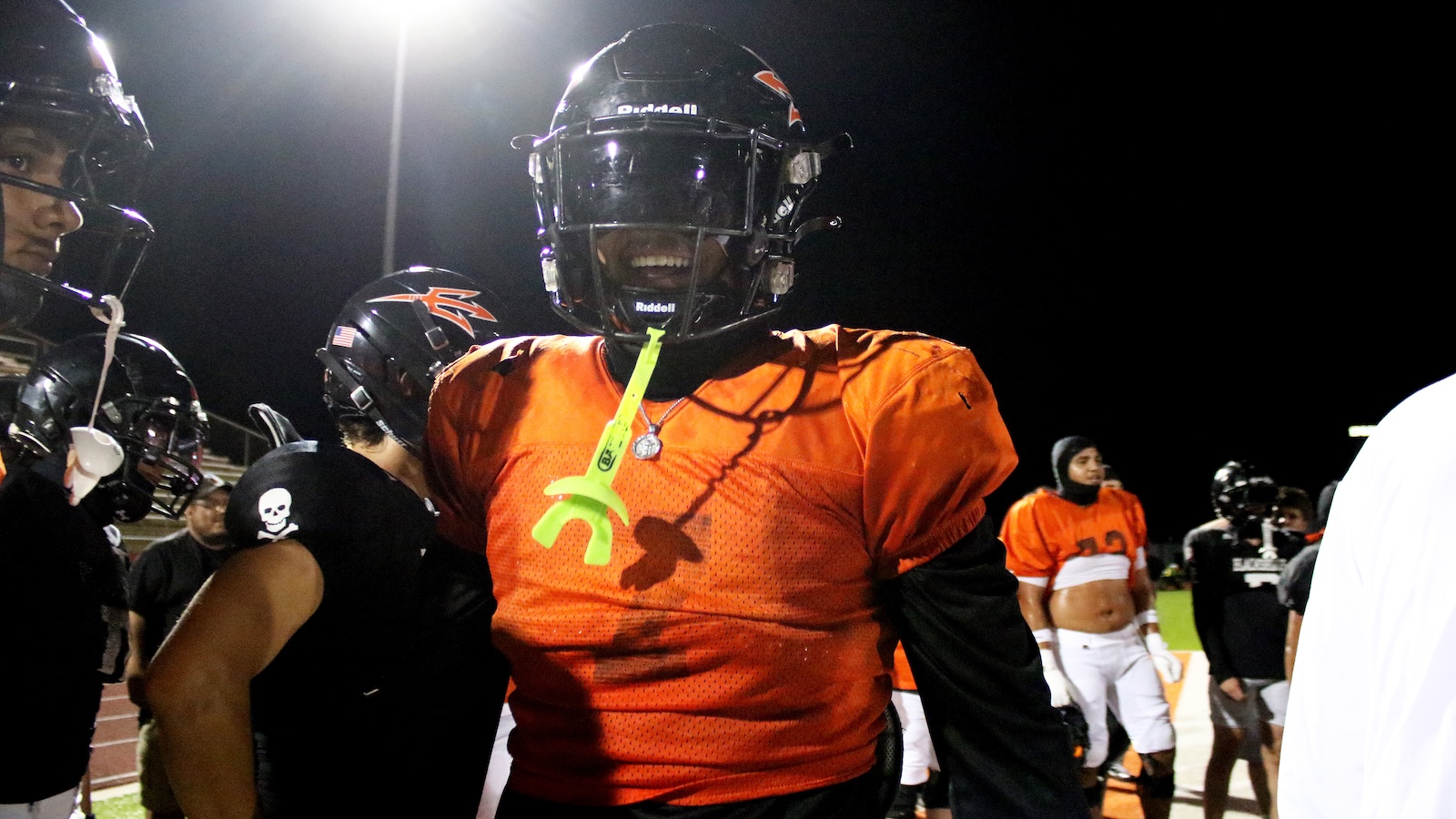 Football player in orange jersey and black helmet on a night field, neon green mouthguard hanging from jersey, teammates in background