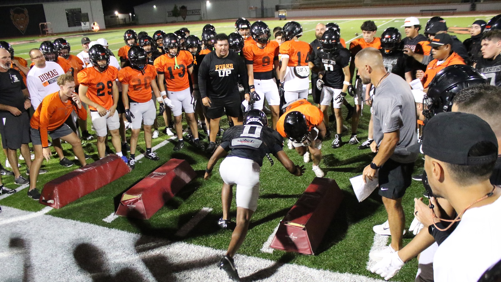 Group of football players in orange and black helmets watch as teammates sprint through padded barriers during a night practice.