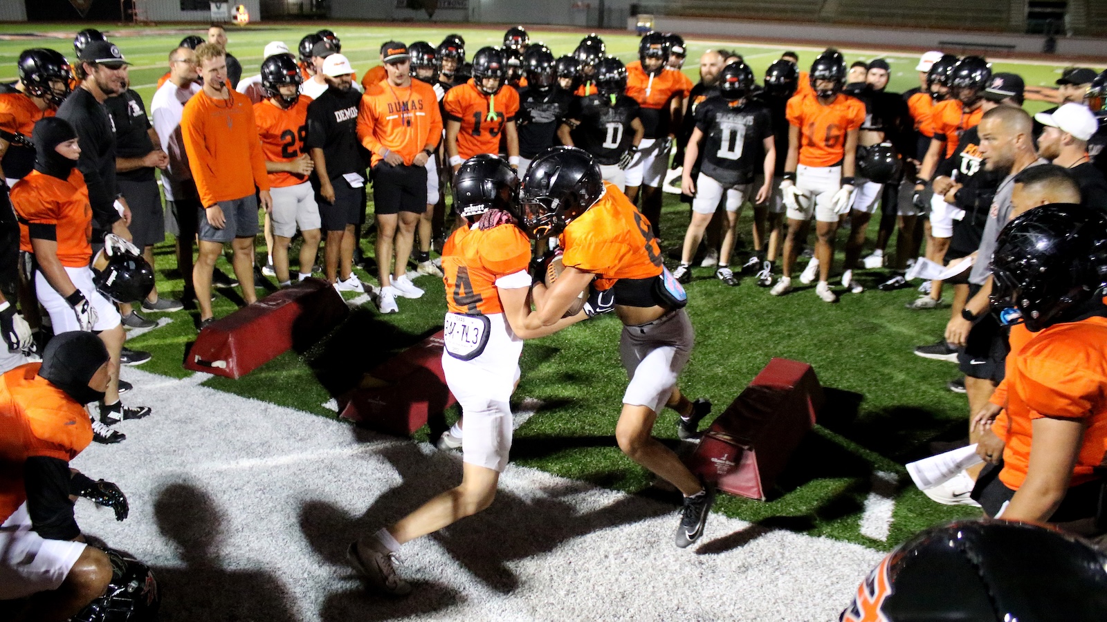 Football players in orange jerseys form a circle around two teammates in a blocking drill on a turf field at night.