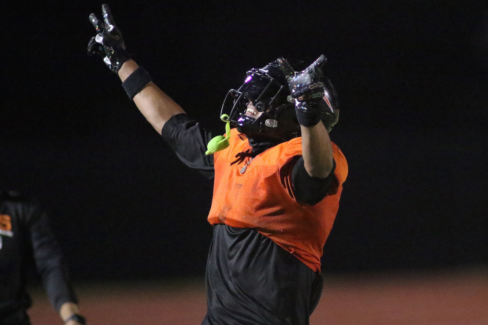 Football player wearing an orange vest and helmet raises arms in celebration on a night field.