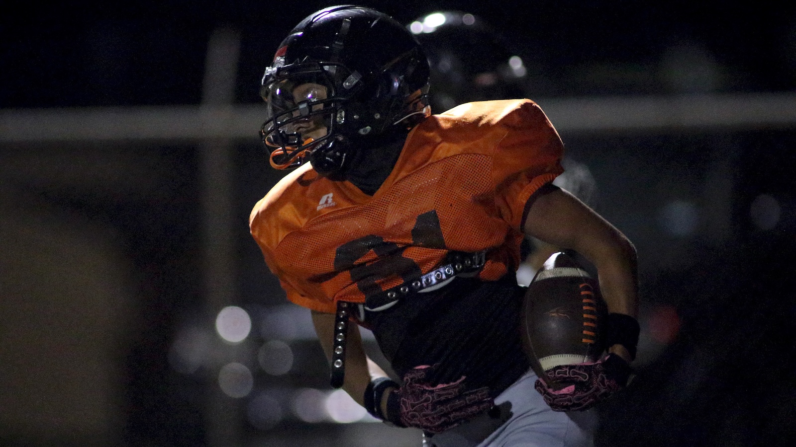 Football player in orange jersey carrying ball, running at night in a dark stadium.