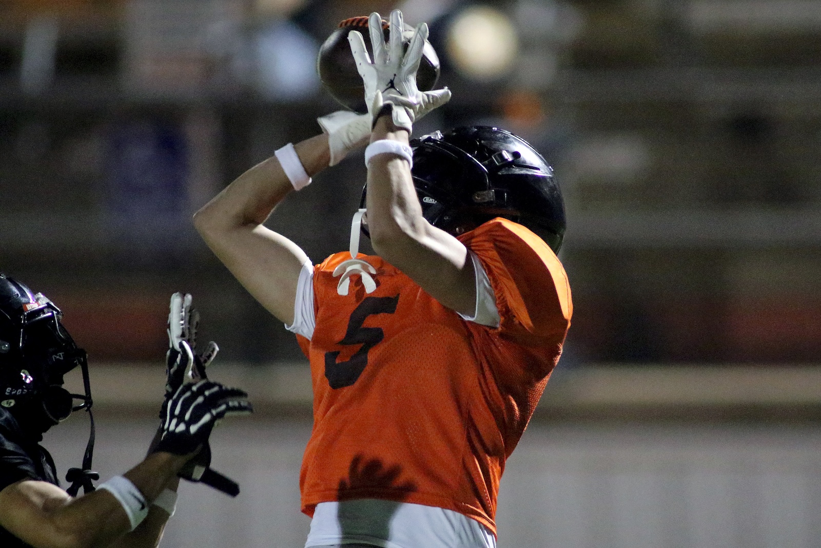 Football players leap for the ball; orange jersey with number 5 reaches up to catch while a defender in black helmet closes in.