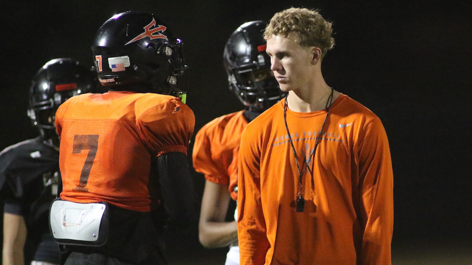 High school football players in orange jerseys standing on a dark field at night; one player with number 7 on his back and a white belt, others in background.