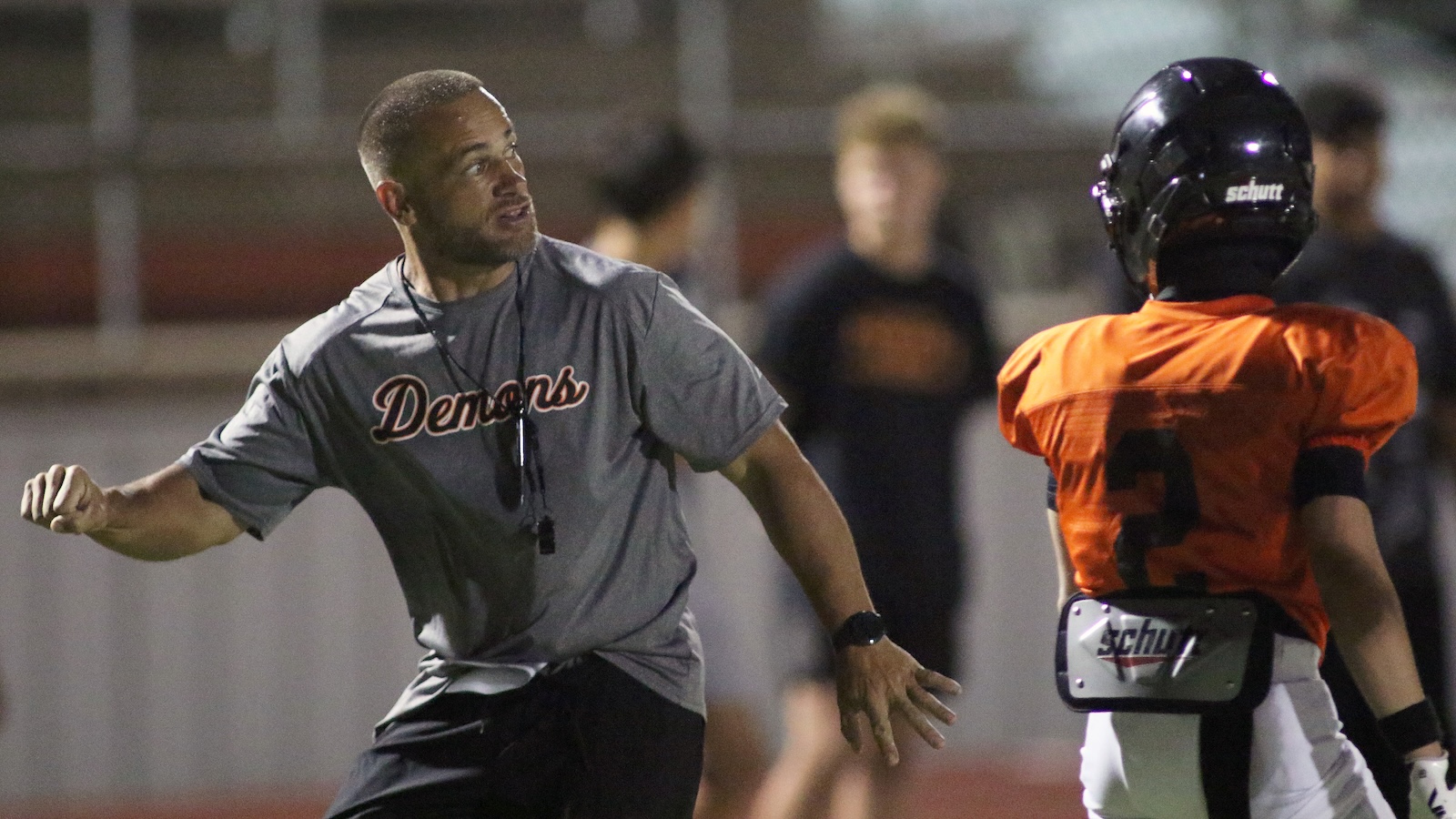Football coach in a gray Demons shirt gestures to players during practice on a field, whistle hanging from neck.